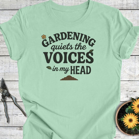 Mint green t-shirt with text 'Gardening quiets the voices in my head' on a wooden surface with gardening tools and flowers.